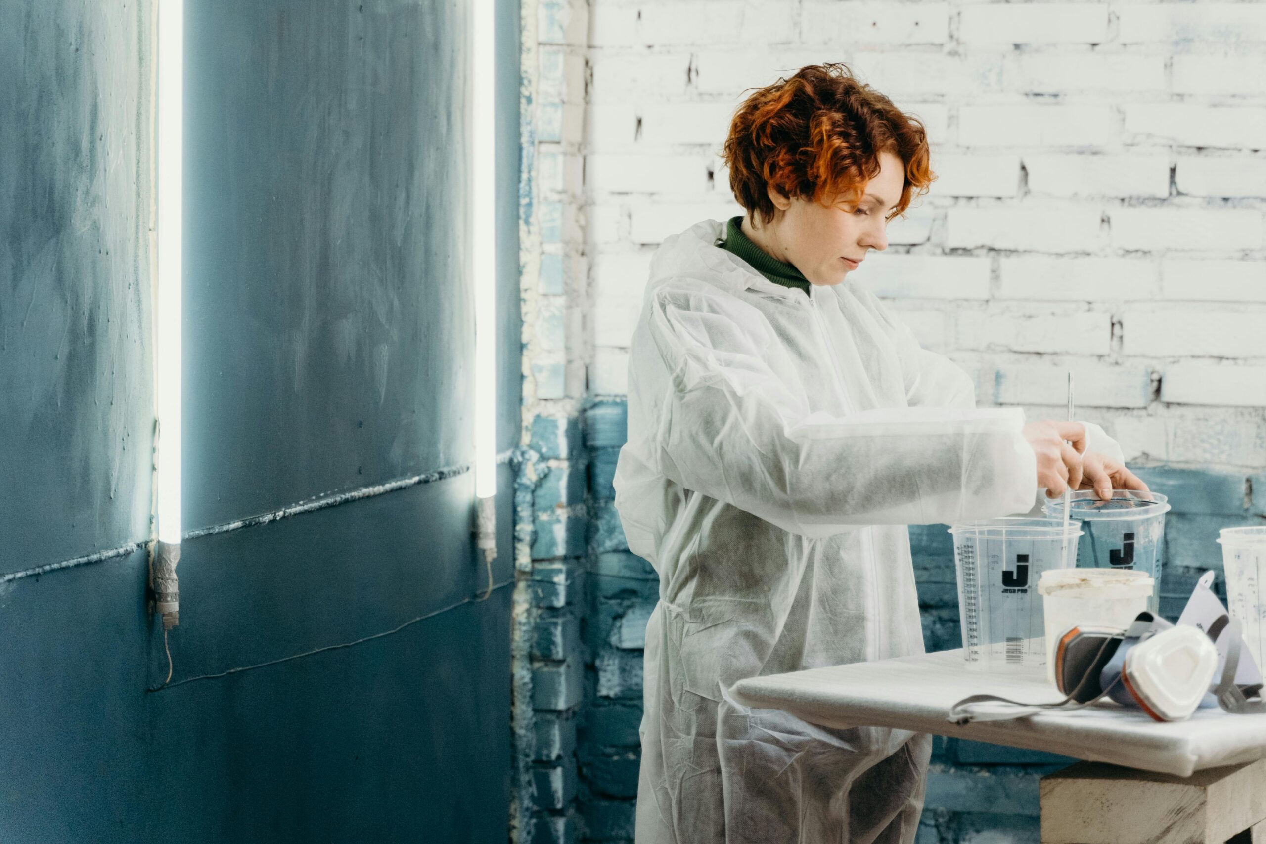 A woman wearing a white protective suit working in a studio environment with a rustic, painted brick wall and fluorescent lighting. She is stirring liquid in a measuring cup, with other cups and a respirator mask placed on a table nearby.