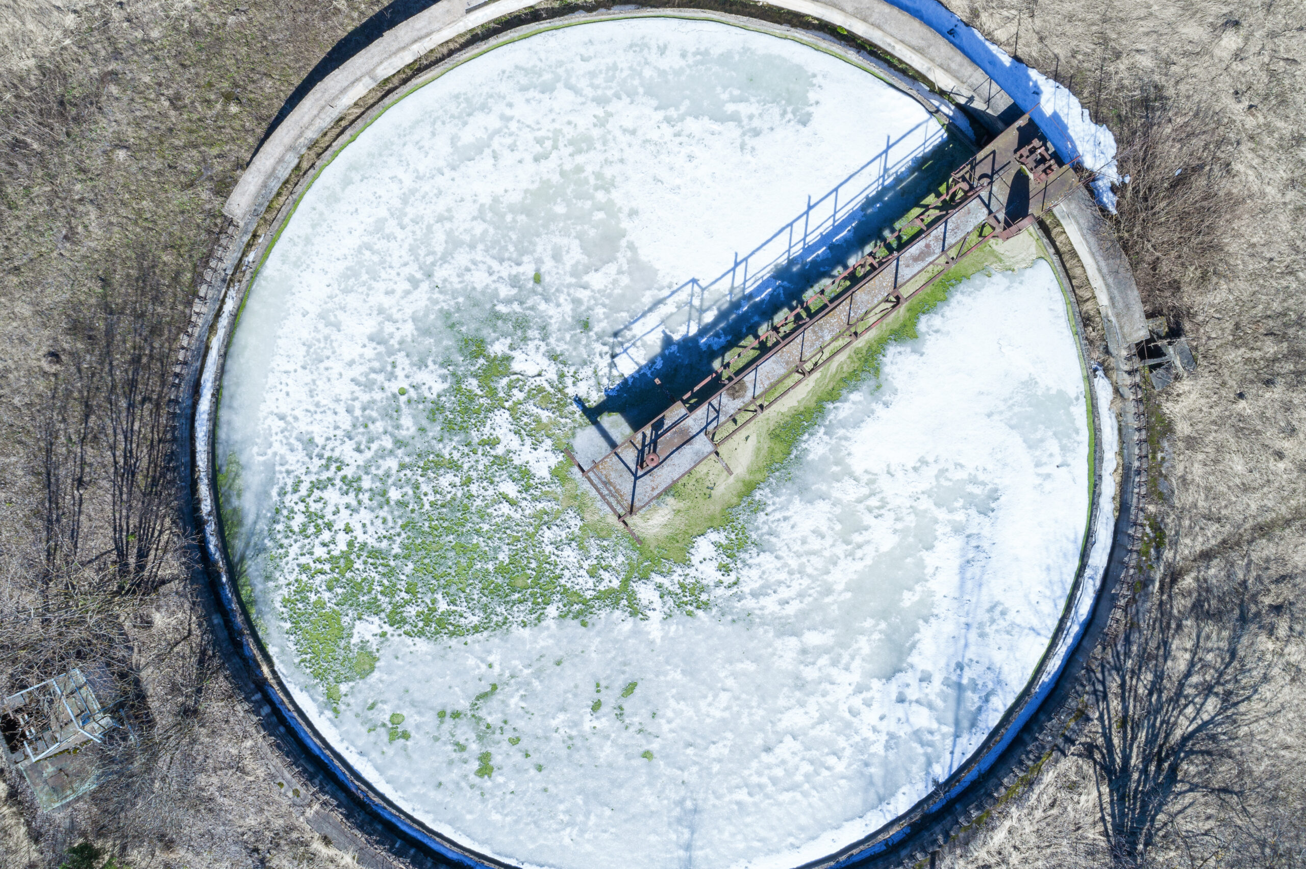 Aerial view of a large circular water treatment or wastewater management facility covered with ice and patches of green algae. The industrial structure is surrounded by dry grassland and features a metal bridge-like structure spanning across its diameter.